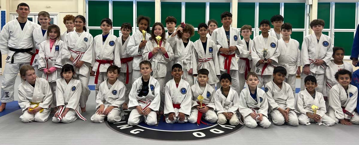 A group of young judo athletes and coaches posing together in the dojo, with several children proudly holding trophies after the Advanced Judo Camp.